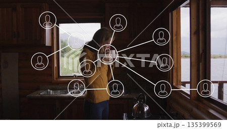 Holding ceramic mug woman gazing through large windows in log cabin kitchen, with network icons 135399569