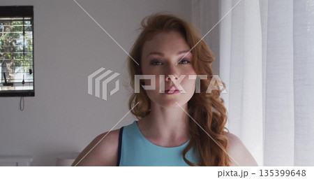 Standing woman wearing sleeveless turquoise top in living area, with blinds, curtains, lamp base Standing woman wearing sleeveless turquoise top in living area, with blinds, curtains, lamp base 135399648