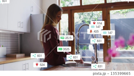 Standing woman wearing burgundy top using high-arc faucet in kitchen, with notification icons 135399665