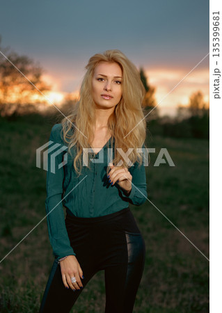 Woman stands in grassy area at sunset, looking at the camera with warm light in the background 135399681