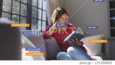 Woman wearing red top, blue jeans, reading book on sofa in living room, with notification icons 135399693