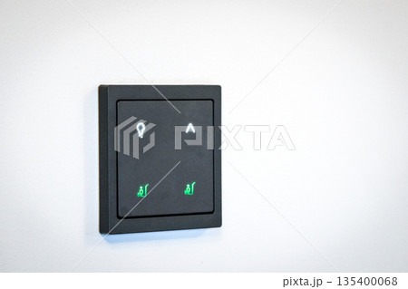 Modern smart home wall control panel with illuminated touch icons on a white wall. Minimalist interior design featuring a black electronic switch for lighting and home automation. Modern smart home wall control panel with illuminated touch icons on a white wall. Minimalist interior design featuring a black electronic switch for lighting and home automation. 135400068
