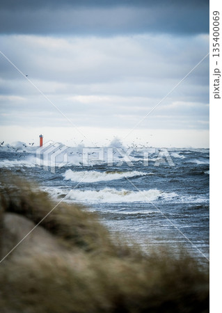 Stormy sea with strong waves crashing under a dramatic cloudy sky. A red navigation beacon stands in the distance while seabirds fly above the rough water. Windy coastal landscape Stormy sea with strong waves crashing under a dramatic cloudy sky. A red navigation beacon stands in the distance while seabirds fly above the rough water. Windy coastal landscape 135400069
