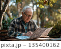 An elderly man with glasses leisurely reads a newspaper at a table 135400162