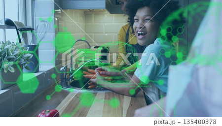 Two women in casual tees washing carrots under faucet in kitchen, with molecular graphics overlay 135400378