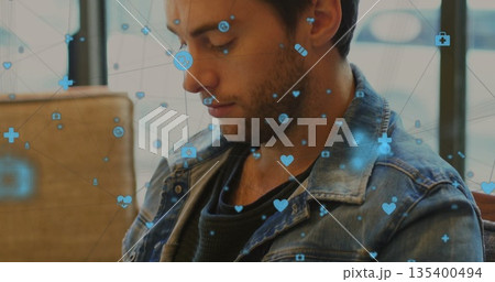 Gazing male wearing denim jacket over dark shirt sitting on seat by window, floating medical icons Gazing male wearing denim jacket over dark shirt sitting on seat by window, floating medical icons 135400494