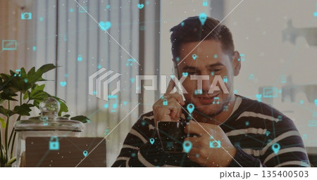 Asian man holding smartphone and scrolling icons on laptop in home office, with plant and jar Asian man holding smartphone and scrolling icons on laptop in home office, with plant and jar 135400503