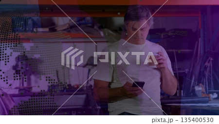 Leaning mature technician holding smartphone and coffee in workshop, shelving and digital overlays 135400530