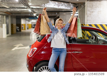 A happy woman raises her arms in triumph after a successful shopping trip, standing next to her red car in a parking garage A happy woman raises her arms in triumph after a successful shopping trip, standing next to her red car in a parking garage 135401509
