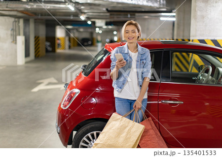 A smiling woman stands next to her red car in a parking garage, holding shopping bags and a phone A smiling woman stands next to her red car in a parking garage, holding shopping bags and a phone 135401533