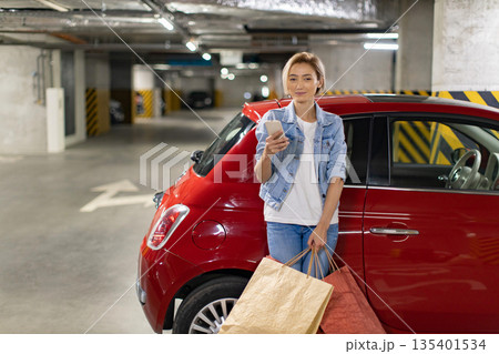A woman stands in a parking garage with shopping bags and a smartphone next to her red car A woman stands in a parking garage with shopping bags and a smartphone next to her red car 135401534