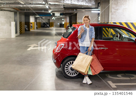 A woman smiles while looking at her phone next to her car after shopping in a parking garage A woman smiles while looking at her phone next to her car after shopping in a parking garage 135401546