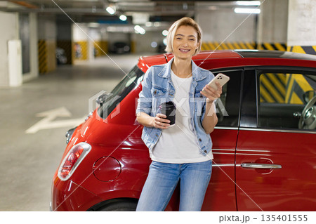 A woman in a denim jacket holds a coffee cup and smartphone while leaning against a red car in a parking garage A woman in a denim jacket holds a coffee cup and smartphone while leaning against a red car in a parking garage 135401555