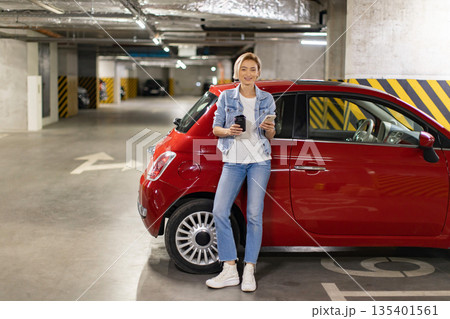 A woman stands next to her red car in a parking garage, holding a coffee cup and a smartphone 135401561