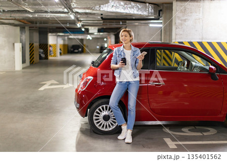 A woman stands next to her red car in an underground parking garage, holding a coffee and her phone 135401562