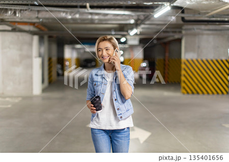 A smiling woman walks through a parking garage while talking on her phone and holding a coffee cup 135401656