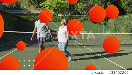 Walking mature adult man and senior woman talking on outdoor tennis court, with tennis rackets 135402202