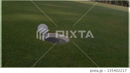 Perched white golf ball balancing on metallic cup edge on putting green, orange markers, copy space Perched white golf ball balancing on metallic cup edge on putting green, orange markers, copy space 135402217