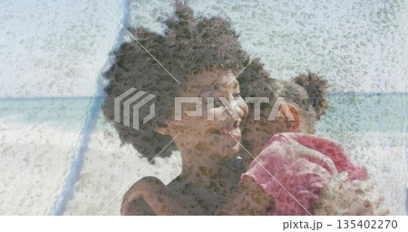 Lifting mother hugging daughter at sandy beach shoreline, displaying curly hair and pink T-shirt 135402270