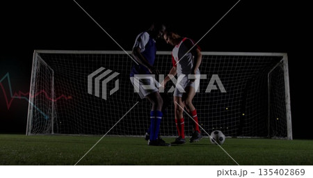 Challenging two opposing soccer players contesting ball on grass pitch under floodlights, goal net 135402869