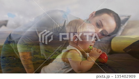 Leaning mother watching daughter mouthing red green teething toy in bedroom, with neutral sheets Leaning mother watching daughter mouthing red green teething toy in bedroom, with neutral sheets 135402909