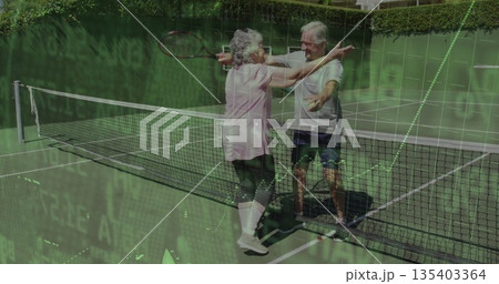 Hugging senior couple in sportswear holding wooden rackets at net on green court, with white lines 135403364