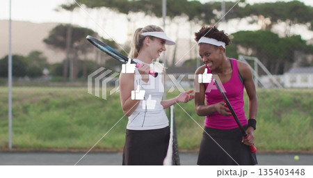 Laughing two women in tennis outfits chatting at net on tennis court, with rackets and ball 135403448