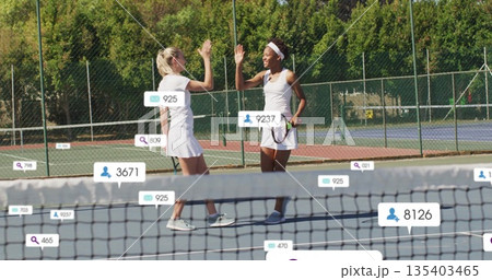 High-fiving tennis teammates in white holding rackets at net on blue court, with floating icons High-fiving tennis teammates in white holding rackets at net on blue court, with floating icons 135403465
