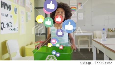 Holding rim of green recycling bin, boy standing in school classroom, with digital thumbs-up icons 135403562