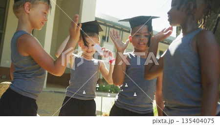 Celebrating four children wearing uniforms in school courtyard with mortarboard hats and diploma Celebrating four children wearing uniforms in school courtyard with mortarboard hats and diploma 135404075