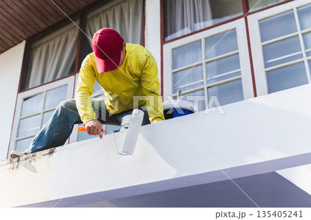 Worker painting an exterior wall with a roller during house maintenance. Worker painting an exterior wall with a roller during house maintenance. 135405241