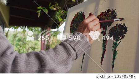 Female artist in grey sweater painting canvas with paintbrush under pergola, with overlay icons 135405497