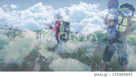 Traversing hikers wearing jackets across field under cloudy sky, carrying poles and packs with pads 135405643