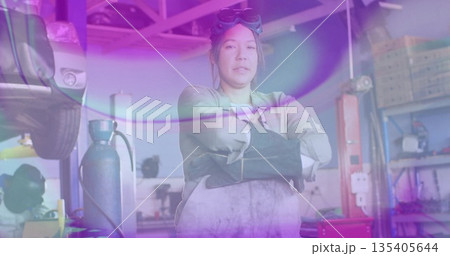 Standing female mechanic wearing gloves and apron crossing arms in garage, displaying vehicle lift 135405644