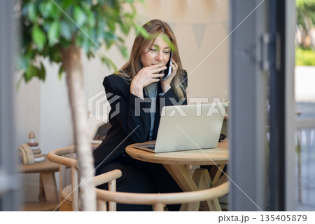 Relaxed woman freelancer enjoying conversation on smartphone while working with laptop in cafe. Relaxed woman freelancer enjoying conversation on smartphone while working with laptop in cafe. 135405879