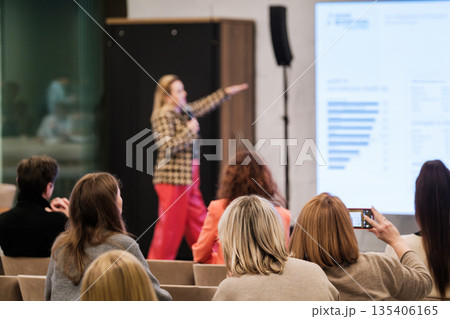 Speaker presents at conference with audience listening and recording during presentation in a modern venue 135406165