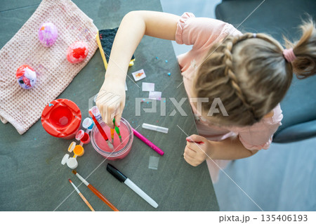 Child painting Easter eggs with brushes and colorful paints at table at home, top view. Creative Easter craft process, festive preparation and hands on activity indoors. Child painting Easter eggs with brushes and colorful paints at table at home, top view. Creative Easter craft process, festive preparation and hands on activity indoors. 135406193