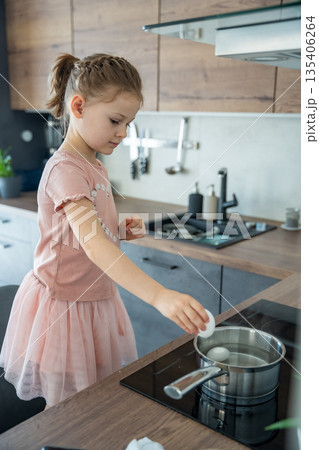 Girl in pink dress standing at the stove preparing to boil Easter eggs. Home holiday preparation and child independence. Girl in pink dress standing at the stove preparing to boil Easter eggs. Home holiday preparation and child independence. 135406264