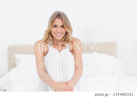 Mid-twenties woman sitting on bed hugging knees in white bedroom wearing pajamas with pillows Mid-twenties woman sitting on bed hugging knees in white bedroom wearing pajamas with pillows 135406331