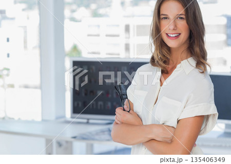 Businesswoman standing confidently holding eyeglasses at desk in bright office with dual monitors 135406349