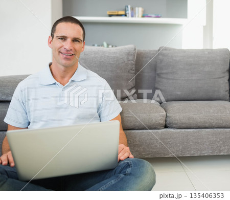 Man sitting on floor holding laptop in living room near grey couch with shelf, copy space Man sitting on floor holding laptop in living room near grey couch with shelf, copy space 135406353