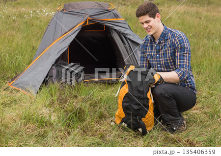 Male camper kneeling at meadow campsite packing hiking backpack by orange dome tent, copy space Male camper kneeling at meadow campsite packing hiking backpack by orange dome tent, copy space 135406359