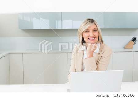 Woman in blazer smiling, using silver laptop on kitchen counter by wooden knife block 135406360