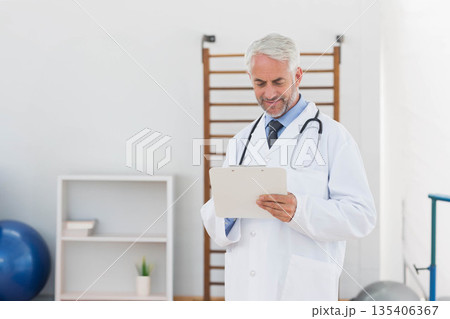 Senior male doctor standing and reading clipboard notes in rehab room with stethoscope, copy space 135406367