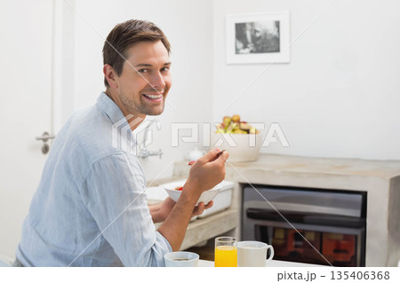 Man in his thirties eating breakfast sitting at kitchen counter holding bowl and spoon 135406368
