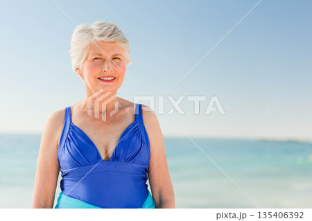Senior woman standing at beach wearing blue one-piece swimsuit and stud earrings Senior woman standing at beach wearing blue one-piece swimsuit and stud earrings 135406392