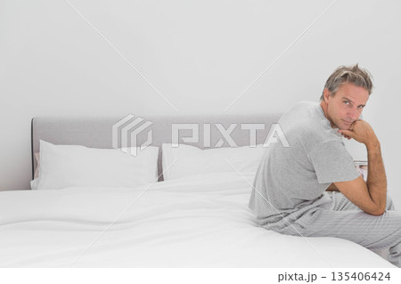 Middle-aged man sitting on bed in bedroom wearing grey T-shirt and striped pajama pants, copy space Middle-aged man sitting on bed in bedroom wearing grey T-shirt and striped pajama pants, copy space 135406424