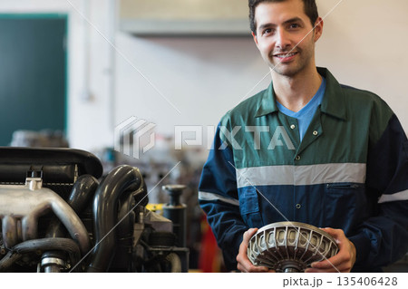 Male mechanic wearing coverall holding converter beside car engine stand in workshop, copy space 135406428
