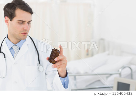 Male doctor holding amber bottle, examining label with stethoscope beside bed in clinic, copy space 135406432
