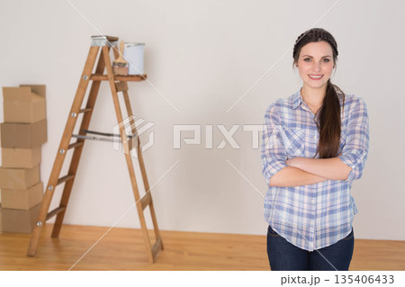 Woman standing with arms crossed in room under renovation with ladder, paint can, boxes, copy space 135406433
